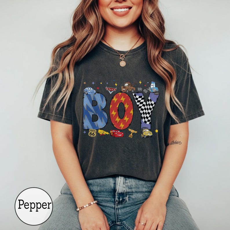 May include: A charcoal gray t-shirt featuring the word BOY in large, patterned letters. The B is blue with lightning bolts, the O is red with yellow lightning, and the Y is black and white checkered. Includes small car character graphics.