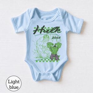 Pu&ograve; includere: Un body per neonati azzurro con una stampa grafica di Hulk e il testo "Hulk 2008" sopra un motivo a scacchi verde e bianco. Il design include una grande illustrazione verde del personaggio e un contorno verde pi&ugrave; piccolo.