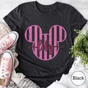 K&ouml;nnte beinhalten: Ein dunkelgraues Kurzarm-T-Shirt mit einer gro&szlig;en Grafik in Form von Mausohren mit rosa und schwarzen vertikalen Streifen. Der Name lily ist in einer dunkelrosa Schreibschrift in der Mitte des Mausohr-Designs aufgedruckt.