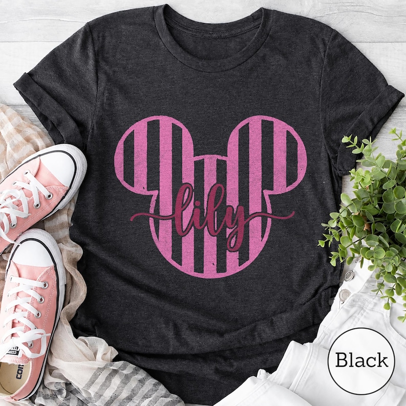 K&ouml;nnte beinhalten: Ein dunkelgraues Kurzarm-T-Shirt mit einer gro&szlig;en Grafik in Form von Mausohren mit rosa und schwarzen vertikalen Streifen. Der Name lily ist in einer dunkelrosa Schreibschrift in der Mitte des Mausohr-Designs aufgedruckt.