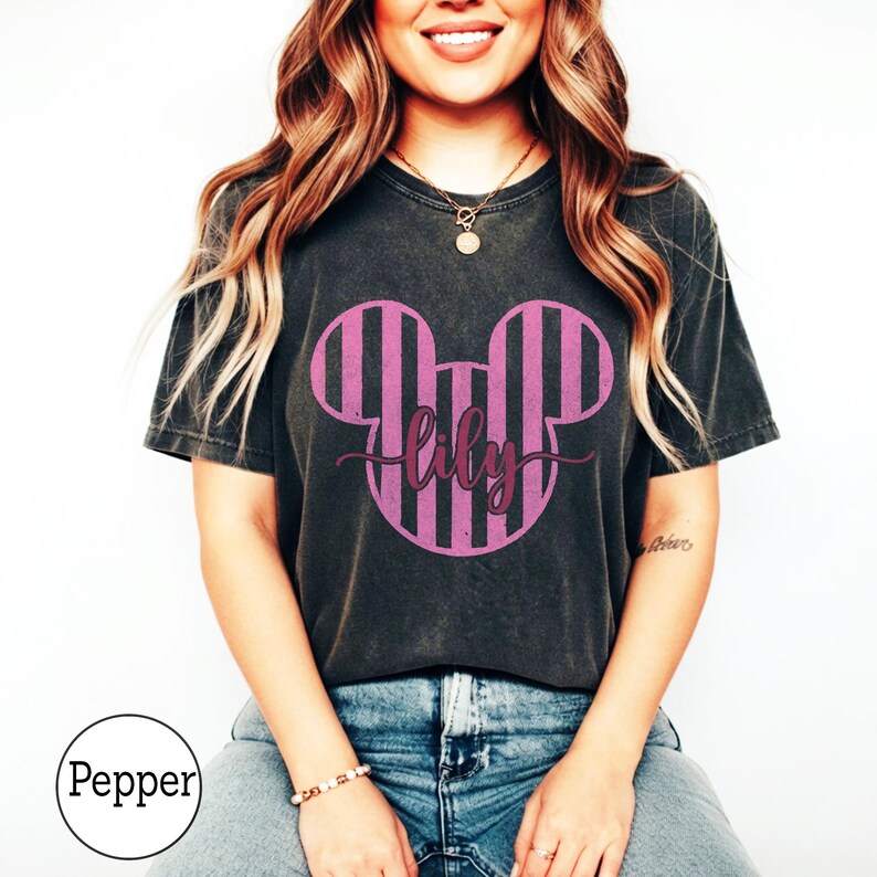 K&ouml;nnte beinhalten: Ein anthrazitfarbenes Kurzarm-T-Shirt mit einer gro&szlig;en Grafik einer Mausohren-Silhouette in Rosa und Schwarz auf der Brust. Der Name lily ist in einer dunkelvioletten Schreibschrift &uuml;ber die Mitte der Grafik gedruckt. Das Shirt hat eine Vintage-Waschung.