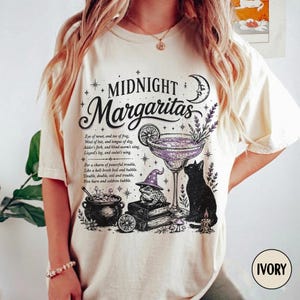 Puede incluir: Una camiseta color marfil con un estampado gráfico negro que incluye el texto Midnight Margaritas, una copa de cóctel, una rana con sombrero morado, un gato negro, un caldero y un poema. El diseño incluye estrellas, luna y lavanda.