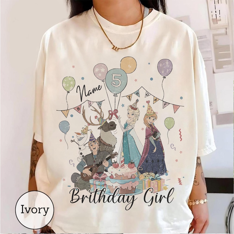 Op de afbeelding: Een ivoorkleurig t-shirt met een grafische print van personages uit Frozen, waaronder Elsa, Anna, Kristoff, Sven en Olaf. Het ontwerp bevat ballonnen, een verjaardagstaart met een kaarsje in de vorm van het cijfer vijf en de tekst Name Birthday Girl.