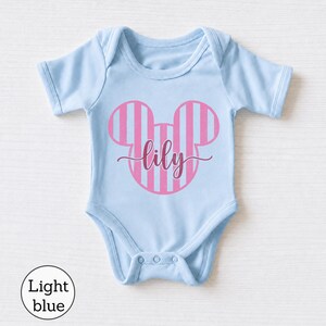 K&ouml;nnte beinhalten: Ein hellblauer Babybody mit einer pink und hellblau gestreiften Mauseohr-Silhouette auf der Brust. Der Name lily ist in einer dunkelpinken Schreibschrift mittig &uuml;ber das Mauseohr-Design auf dem Kleidungsst&uuml;ck gedruckt.