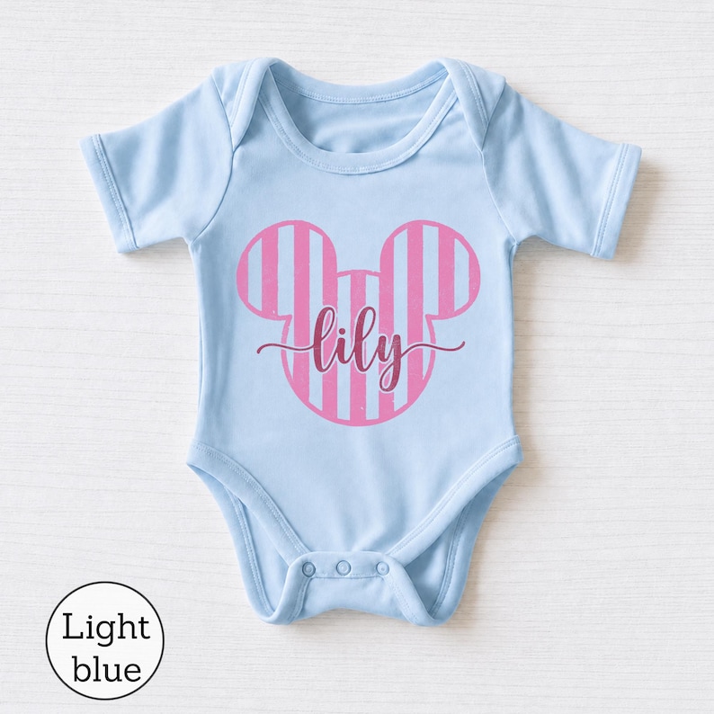 K&ouml;nnte beinhalten: Ein hellblauer Babybody mit einer pink und hellblau gestreiften Mauseohr-Silhouette auf der Brust. Der Name lily ist in einer dunkelpinken Schreibschrift mittig &uuml;ber das Mauseohr-Design auf dem Kleidungsst&uuml;ck gedruckt.