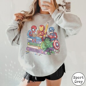 Puede incluir: Una sudadera gris claro de cuello redondo con un gr&aacute;fico de superh&eacute;roes de dibujos animados, incluyendo a Spider-Man, Iron Man, Captain America y Hulk, sentados sobre una pila de libros. Los libros dicen Super Physics, World History y Avengers Assemble.