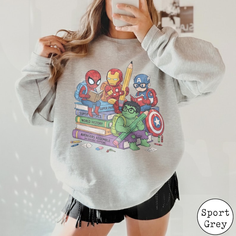 Puede incluir: Una sudadera gris claro de cuello redondo con un gr&aacute;fico de superh&eacute;roes de dibujos animados, incluyendo a Spider-Man, Iron Man, Captain America y Hulk, sentados sobre una pila de libros. Los libros dicen Super Physics, World History y Avengers Assemble.