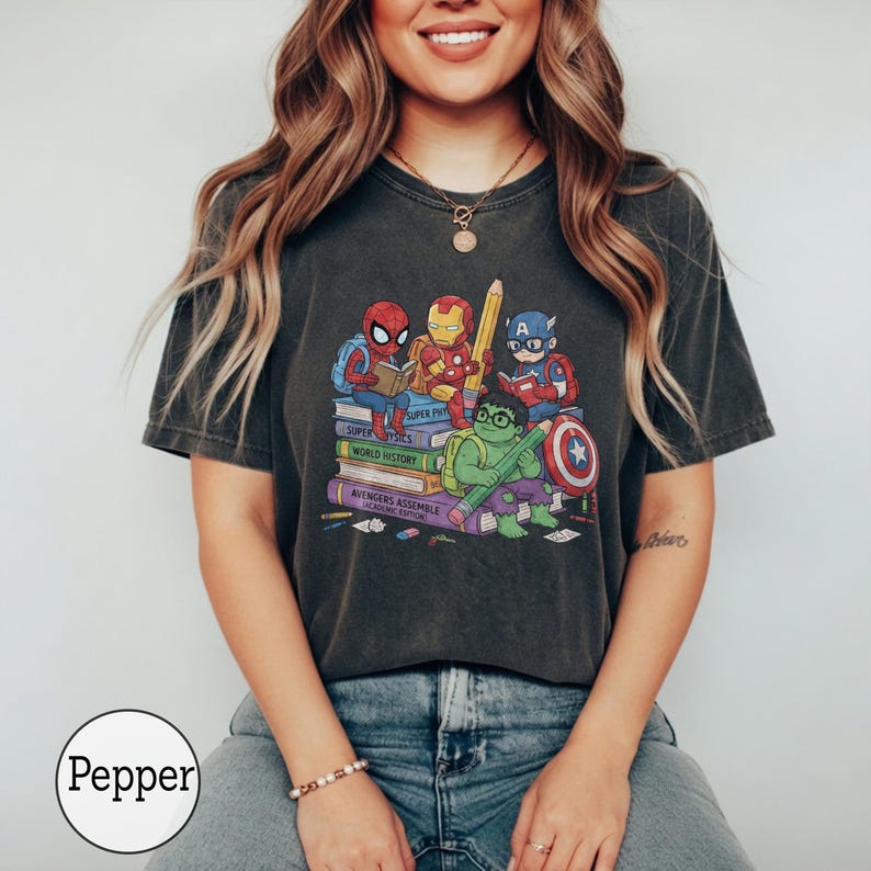 Puede incluir: Una camiseta gris carb&oacute;n de manga corta con un gr&aacute;fico de superh&eacute;roes de dibujos animados sentados sobre una pila de libros. Los libros est&aacute;n etiquetados como Super Physics, World History y Avengers Assemble Academic Edition. Los personajes incluyen a Spider-Man, Iron Man, Captain America y Hulk.