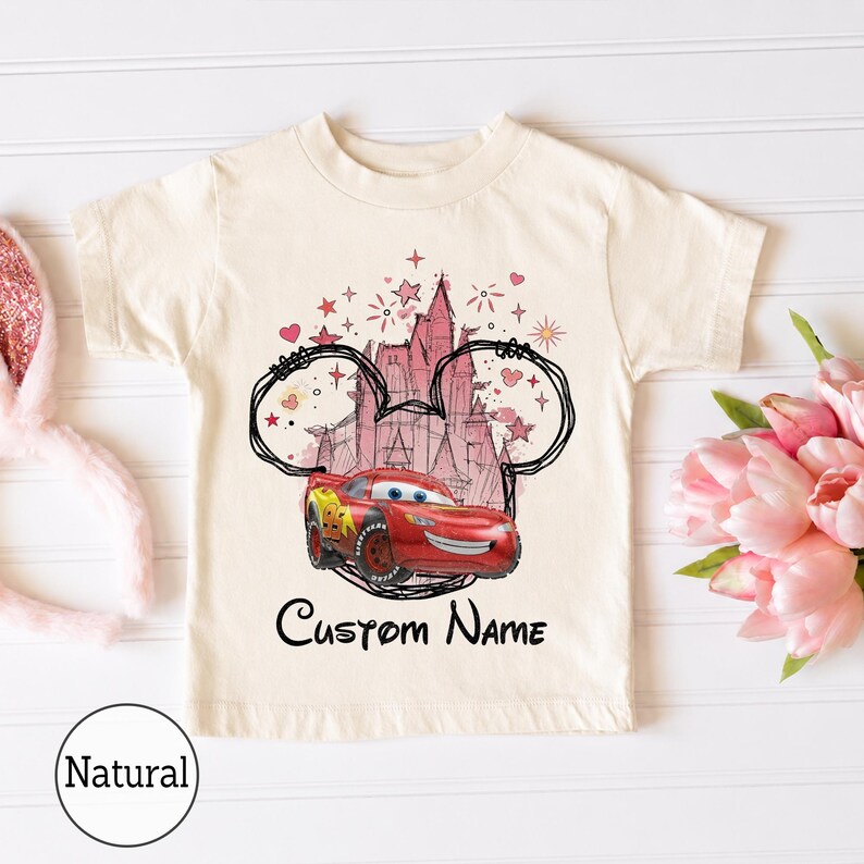 K&ouml;nnte beinhalten: Ein naturfarbenes Kurzarm-T-Shirt mit einem Grafikdruck eines roten Rennwagens vor der Silhouette eines rosa Schlosses, umrahmt von einer schwarzen Umrisslinie von Mausohren, kleinen Sternen, Herzen und dem Text Custom Name darunter.