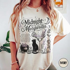 May include: An ivory t-shirt featuring a black graphic print with the text Midnight Margaritas. The design includes a black cat, a cocktail glass, a cauldron, lavender sprigs, a crescent moon, and small text detailing a potion recipe.