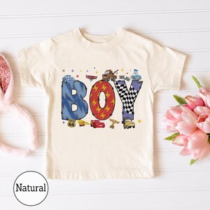 May include: A natural-colored short-sleeve t-shirt featuring a graphic print of the word BOY. The letters are decorated with blue lightning bolts, red lightning patterns, and black and white checkered flags, surrounded by various cartoon car characters.