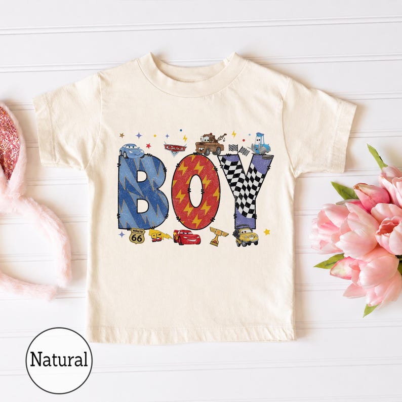 May include: A natural-colored short-sleeve t-shirt featuring a graphic print of the word BOY. The letters are decorated with blue lightning bolts, red lightning patterns, and black and white checkered flags, surrounded by various cartoon car characters.