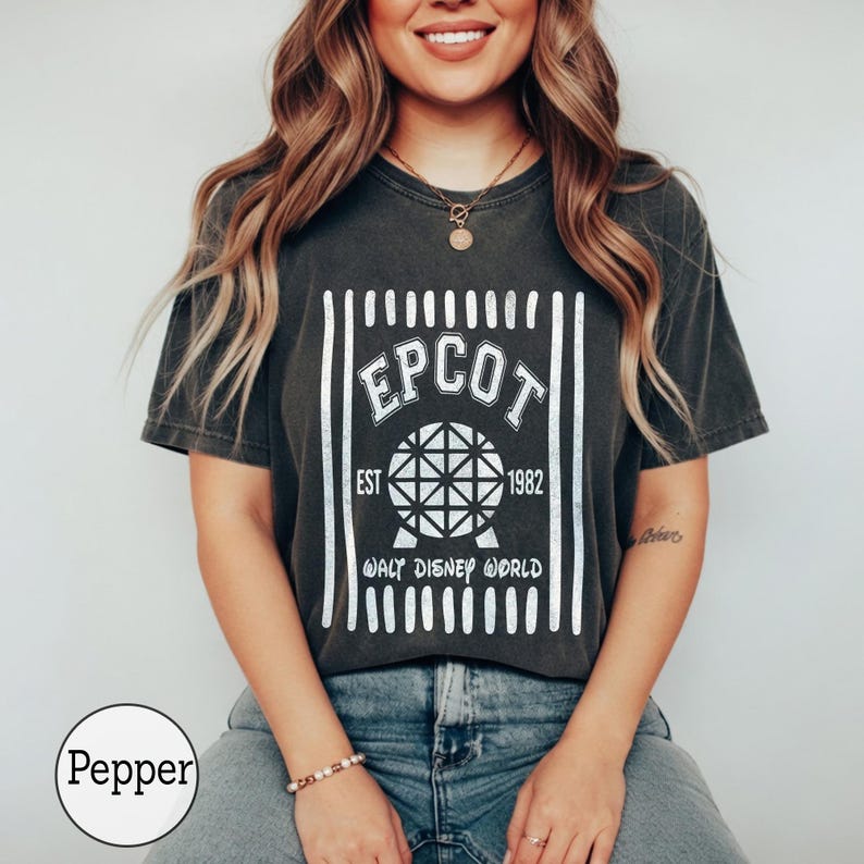 Pu&ograve; includere: Una t-shirt a maniche corte grigio antracite con una stampa grafica bianca. Il design include il testo EPCOT, EST 1982 e WALT DISNEY WORLD, centrato attorno a un'icona a sfera geometrica, il tutto incorniciato da linee verticali bianche.