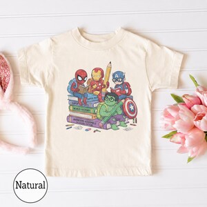 Puede incluir: Una camiseta de manga corta de color natural con un estampado gr&aacute;fico de superh&eacute;roes de dibujos animados, incluidos Spider-Man, Iron Man, Captain America y Hulk, sentados sobre una pila de libros titulados Super Physics, World History y Avengers Assemble.