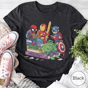 Puede incluir: Una camiseta gris carb&oacute;n oscuro con un estampado gr&aacute;fico de superh&eacute;roes de dibujos animados, Spider-Man, Iron Man, Captain America y Hulk, sentados sobre una pila de libros. Los libros est&aacute;n etiquetados como Super Physics, World History y Avengers Assemble.