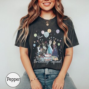 Op de afbeelding: Een antracietgrijs T-shirt met korte mouwen en een grafische print van animatiefiguren, ballonnen en een verjaardagstaart. De tekst luidt "Name", "5" en "Brithday Squad". Het shirt heeft een vintage gewassen afwerking en een ronde hals.