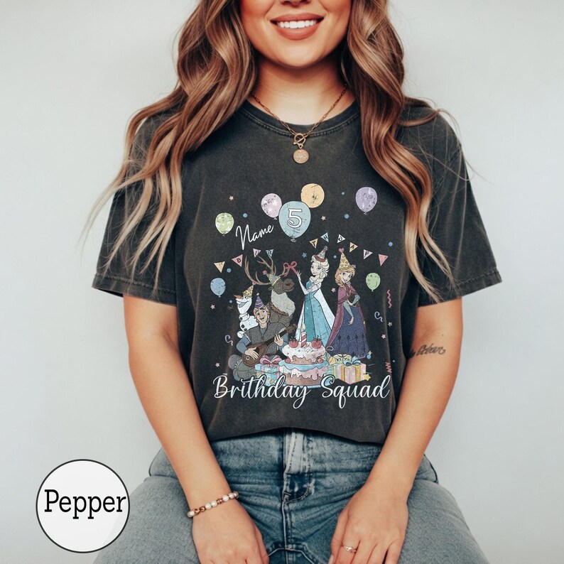 Op de afbeelding: Een antracietgrijs T-shirt met korte mouwen en een grafische print van animatiefiguren, ballonnen en een verjaardagstaart. De tekst luidt "Name", "5" en "Brithday Squad". Het shirt heeft een vintage gewassen afwerking en een ronde hals.