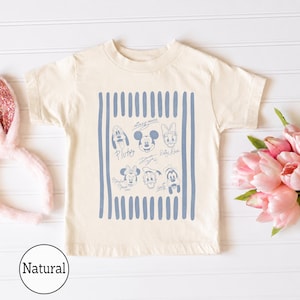 May include: A natural-colored short-sleeve t-shirt featuring a blue graphic print of Disney character faces and signatures, including Mickey Mouse, Minnie Mouse, Donald Duck, Daisy Duck, Pluto, and Goofy, framed by vertical blue stripes.