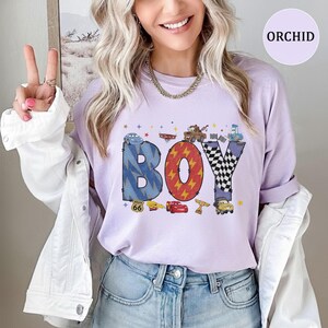 May include: A light purple t-shirt featuring the word BOY in large, patterned letters. The B is blue with lightning bolts, the O is red with yellow lightning, and the Y is black and white checkered. Characters from the movie Cars are included.