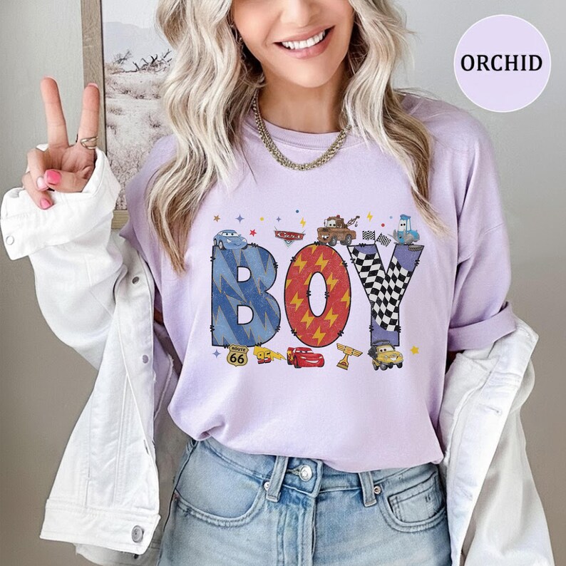 May include: A light purple t-shirt featuring the word BOY in large, patterned letters. The B is blue with lightning bolts, the O is red with yellow lightning, and the Y is black and white checkered. Characters from the movie Cars are included.