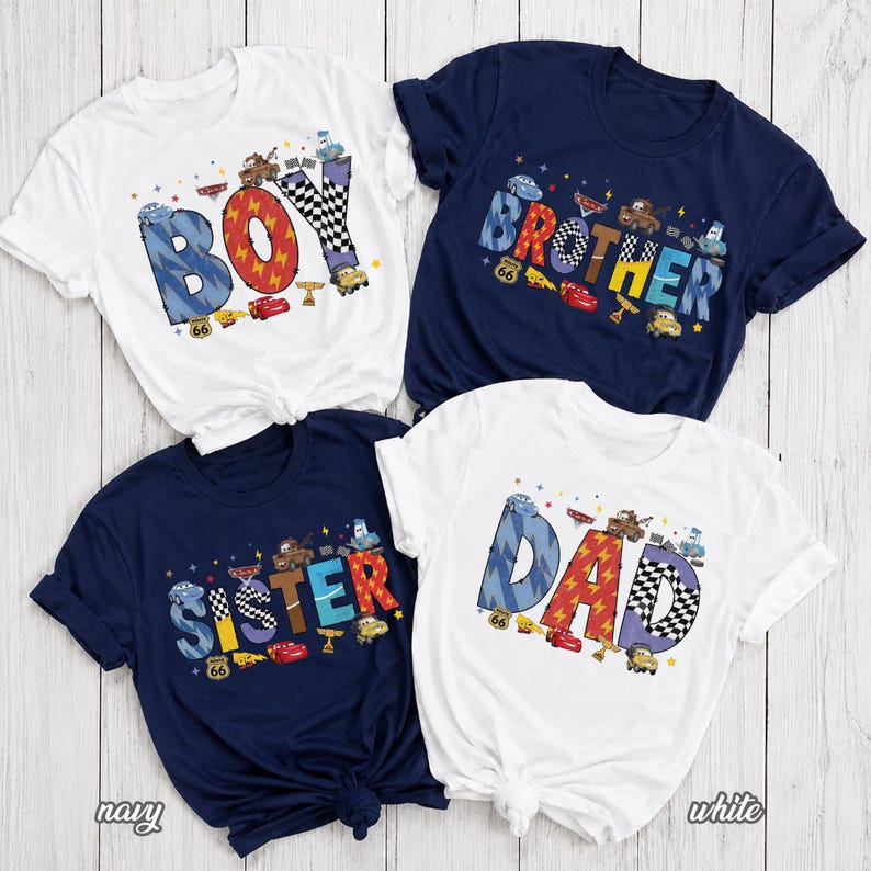 May include: Four short-sleeve t-shirts in white and navy blue. Each shirt features a large word printed in a colorful, cartoon-style font decorated with race car graphics and checkered patterns. The words displayed are BOY, BROTHER, SISTER, and DAD.