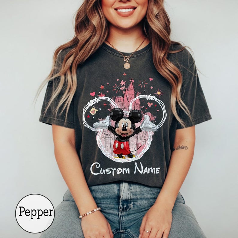 May include: A charcoal gray short-sleeve t-shirt featuring a graphic of a Mickey Mouse balloon character centered within a white hand-drawn ear outline. The design includes a pink castle silhouette, stars, hearts, and the text Custom Name in white.
