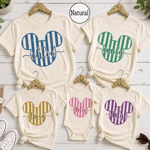 K&ouml;nnte beinhalten: F&uuml;nf cremefarbene T-Shirts und ein Body auf goldenen Kleiderb&uuml;geln, jeweils mit einer gestreiften Mickey-Maus-Kopf-Silhouette. Die Designs enthalten die Namen chloe in Blau, emily in Gr&uuml;n, daisy in Gold sowie lily in Pink und Lila. Text Natural sichtbar.