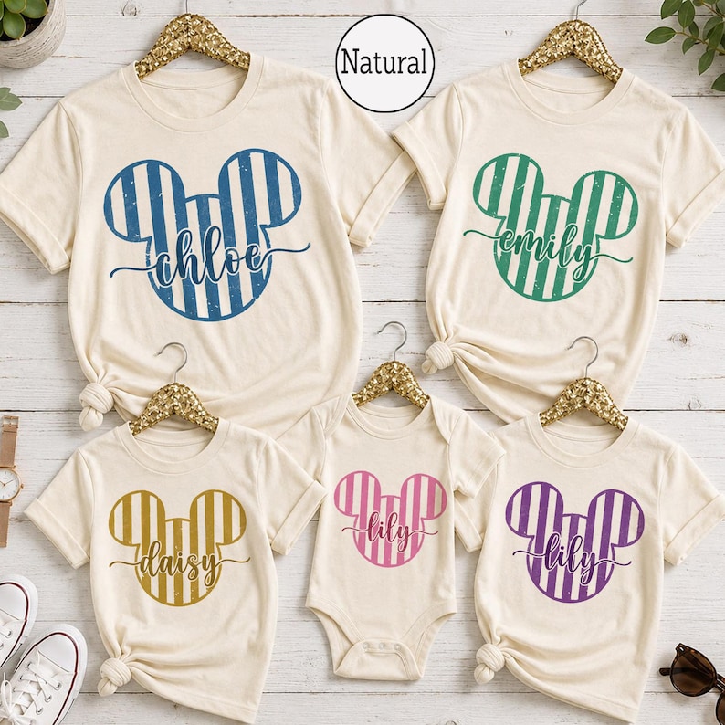 K&ouml;nnte beinhalten: F&uuml;nf cremefarbene T-Shirts und ein Body auf goldenen Kleiderb&uuml;geln, jeweils mit einer gestreiften Mickey-Maus-Kopf-Silhouette. Die Designs enthalten die Namen chloe in Blau, emily in Gr&uuml;n, daisy in Gold sowie lily in Pink und Lila. Text Natural sichtbar.