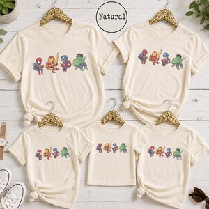 May include: Five natural-colored t-shirts of varying sizes, each featuring a graphic print of four cartoon superheroes: Spider-Man, Iron Man, Captain America, and Hulk. Each character is depicted with school supplies like books and pencils.