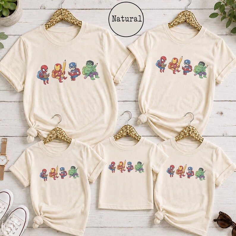 May include: Five natural-colored t-shirts of varying sizes, each featuring a graphic print of four cartoon superheroes: Spider-Man, Iron Man, Captain America, and Hulk. Each character is depicted with school supplies like books and pencils.