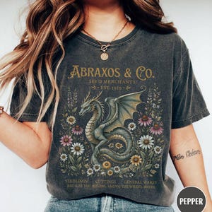 Puede incluir: Una camiseta gris carbón con un gráfico de un dragón rodeado de flores silvestres. El texto dice ABRAXOS & CO. SEED MERCHANTS EST. 1910 y SEEDLINGS CUTTINGS GENERAL WARES BECAUSE YOU BELONG AMONG THE WILDFLOWERS en letras doradas.