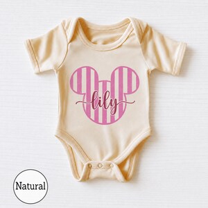 K&ouml;nnte beinhalten: Ein naturfarbener Kurzarm-Babybody mit einer Grafik in Form von Mausohren mit rosa-wei&szlig;en Streifen auf der Brust. Der Name lily ist in einer dunkelrosa Schreibschrift in der Mitte des Mausohren-Motivs aufgedruckt.