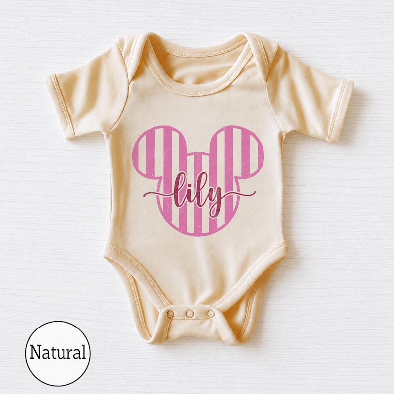 K&ouml;nnte beinhalten: Ein naturfarbener Kurzarm-Babybody mit einer Grafik in Form von Mausohren mit rosa-wei&szlig;en Streifen auf der Brust. Der Name lily ist in einer dunkelrosa Schreibschrift in der Mitte des Mausohren-Motivs aufgedruckt.