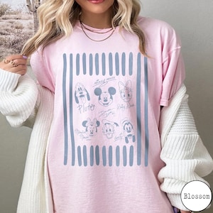 May include: A light pink t-shirt featuring a central graphic of blue vertical stripes and line-art illustrations of cartoon characters including Mickey Mouse, Minnie Mouse, Donald Duck, Daisy Duck, Pluto, and Goofy, along with their printed signatures.