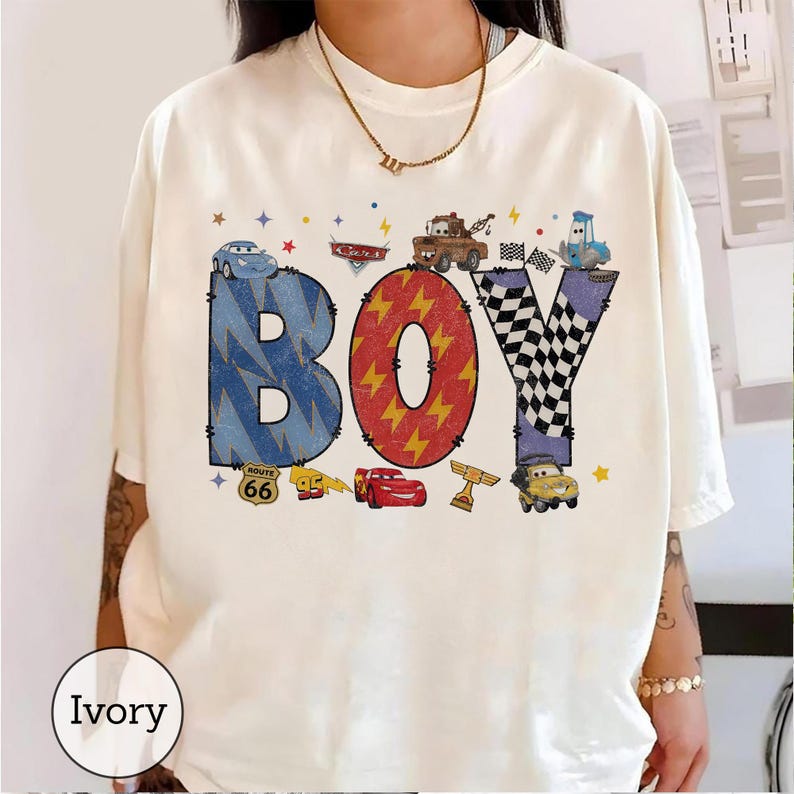 May include: An ivory t-shirt featuring the word BOY in large, colorful letters. The B is blue with lightning bolts, the O is red with yellow lightning, and the Y is black and white checkered. Various cartoon cars and racing icons surround the text.