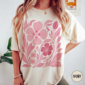 Może przedstawiać: T-shirt z krótkim rękawem w kolorze kości słoniowej z dużym różowym wzorem kwiatowym. Nadruk zawiera różne kwiaty, liście i małe wstążki świadomości, z tekstem A CURE WORTH FIGHTING FOR wydrukowanym w łuku po prawej stronie.
