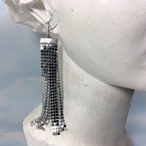 May include: Silver chain earrings with a fringe design. The earrings are dangling from a mannequin's ear.