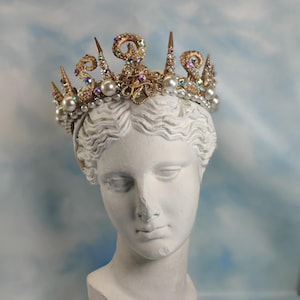 May include: A gold-colored crown with pearls and iridescent crystals sits atop a white bust of a woman. The crown has ornate details and pointed accents. The background is a light blue sky.