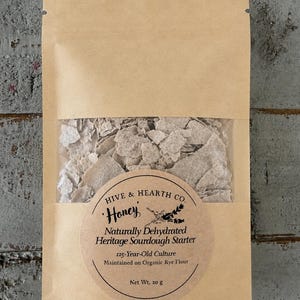 May include: A tan paper pouch containing dehydrated sourdough starter flakes. A circular label reads: Hive & Hearth Co. Honey, Naturally Dehydrated Heritage Sourdough Starter, 125-Year-Old Culture, Maintained on Organic Rye Flour, Net Wt. 20 g.