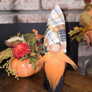 May include: A small gnome figurine with a black and white plaid hat, orange beard, and black body. The gnome is standing on a wooden surface.