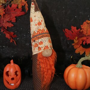 Puede incluir: Un gnomo artesanal con barba naranja y un gorro con calabazas y la palabra "PUMPKINS". Una calabaza naranja tallada y una calabaza redonda naranja completan la escena, evocando el otoño y Halloween.