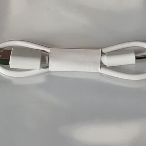 Könnte beinhalten: Ein weißes USB-Ladekabel, das zu einer ovalen Form aufgewickelt ist. Das Kabel verfügt an einem Ende über einen standardmäßigen rechteckigen USB-A-Anschluss und am anderen Ende über einen kleineren, abgerundeten USB-C-Anschluss. Ein weißes Papierband fixiert die Mitte.