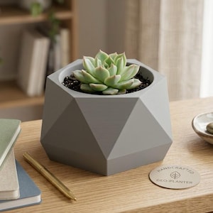 Puede incluir: Una maceta geométrica de color gris claro con una superficie facetada que contiene una planta suculenta verde. Junto a la maceta hay una etiqueta redonda de color beige con el texto HANDCRAFTED GEO-PLANTER y un icono de hoja en marrón.