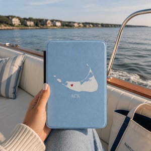 May include: A light blue e-reader case featuring a white silhouette map of Nantucket island with a small red heart icon placed on the map and the text ACK printed in white below the map, all centered on the front cover of the protective case.