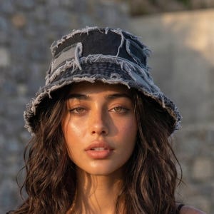 Può includere: Un cappello da pescatore grigio antracite scuro caratterizzato da un design sfrangiato con cuciture a vista e toppe in tessuto denim grezzo. Il cappello presenta una corona strutturata e una tesa spiovente, con un'estetica testurizzata e vissuta.