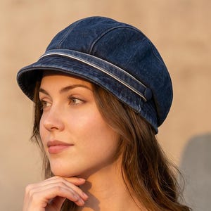 May include: A dark blue denim newsboy cap featuring a structured crown with multiple stitched panels and a short, curved brim. The cap is accented by a horizontal white and blue striped band detail that wraps around the base of the crown.