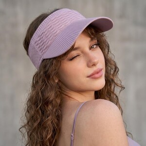 May include: A light purple visor featuring a textured, cable-knit pattern on the front panel and a smooth, ribbed brim. The visor is constructed from a knit fabric with a visible mesh-like texture on the side panels, providing a structured fit.
