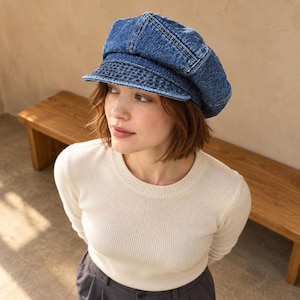 May include: A blue denim newsboy cap featuring a structured crown with multiple stitched panels and a short, stiff brim. The fabric has a classic medium-wash texture with visible seams, providing a rounded shape that sits comfortably on the head.