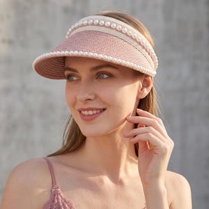 Peut inclure: Une visière en paille rose poudré dotée d'un large bord structuré et d'un bandeau en tissu beige. La visière est ornée de deux rangées de fausses perles blanches sphériques qui bordent les bords supérieur et inférieur du bandeau texturé.