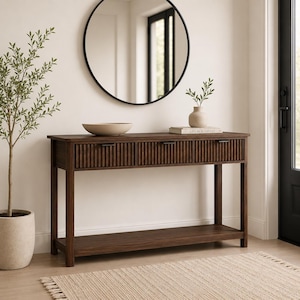 May include: A dark brown wooden console table featuring three drawers with vertical fluted detailing and slim black metal handles. The table has four straight legs and a lower open shelf for storage, providing a functional surface for home decor.
